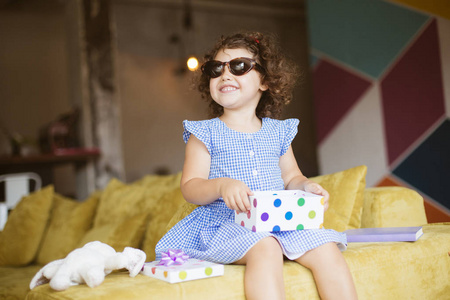 可爱的小微笑的女孩与深色卷曲的头发在蓝色礼服和太阳镜与礼物盒在膝盖愉快地看一边沙发在家里