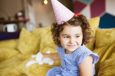 美丽的小微笑的女孩与深色卷曲的头发在蓝色礼服和生日帽子愉快地看一边与兔子玩具在手在沙发在家