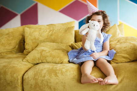 美丽的女孩与深色卷曲的头发在蓝色礼服和生日帽子愉快地显示兔子玩具在照相机在家庭沙发上