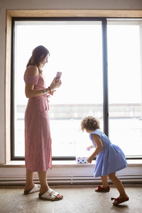 漂亮的女人在礼服愉快地拍摄她的小可爱的女儿在手机上的照片背景大窗口