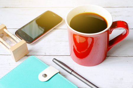 在白色木质背景下, 带有笔记本钢笔沙漏和智能手机的红色杯中的黑咖啡的选择性聚焦和顶级视图