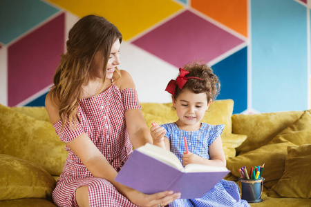 美丽的年轻母亲与她的小可爱的女儿愉快地画在着色书与毛毡笔花时间一起在沙发上在家