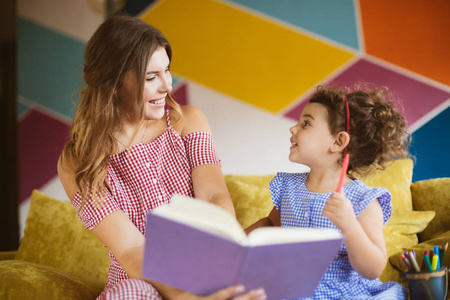 美丽的微笑的母亲与她的小可爱的女儿愉快地看彼此在沙发上一起读儿童书在家