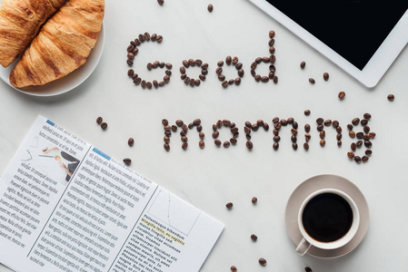 咖啡杯的顶部视图和早晨用牛角面包报纸和片剂制成的白色表面的咖啡豆刻字