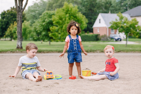 三可爱的白种人和拉美裔儿童的肖像, 孩子们坐在沙盒里玩塑料五颜六色的玩具。小女孩和男孩朋友一起在操场上玩得开心