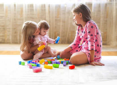 三个金发碧眼的小女孩在室内玩着许多五颜六色的玩具积木。与建筑和创造乐趣的姐妹