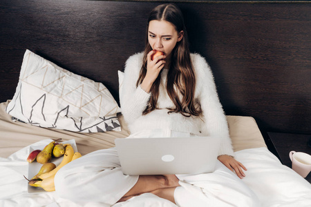 长头发的年轻女孩坐在床上看电视节目在她的笔记本电脑和吃苹果