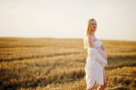 金色的孕妇在花圈领域在白色内衣衣服日落。怀孕的快乐时刻