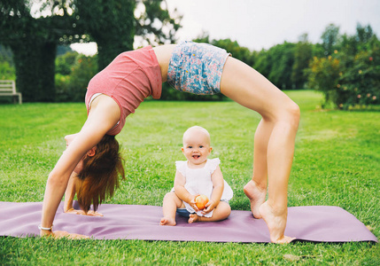 在夏日公园里和婴儿一起做瑜伽的年轻女子。美丽的母亲和女儿就像一只小佛像, 在大自然的青草上锻炼。家庭户外。父母与孩子一起度过时光