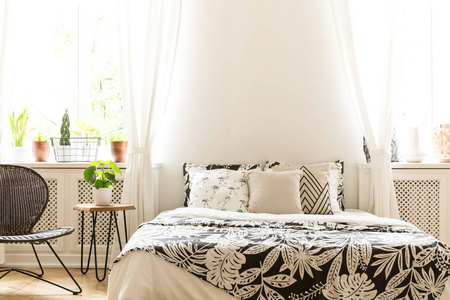 在阳光明媚的卧室内的床上, 黑色和白色的叶子图案的特写。一个藤和金属椅子和一个在房间里的植物的凳子。真实照片