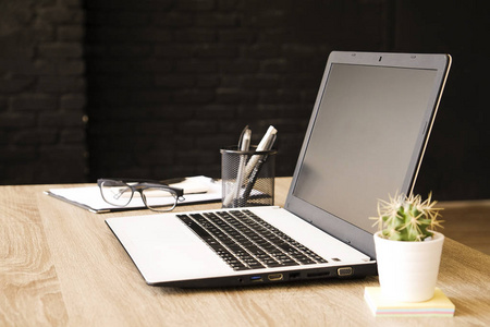 白色笔记本电脑和折叠眼睛眼镜在阁楼式办公室与黑色砖墙的木桌上.