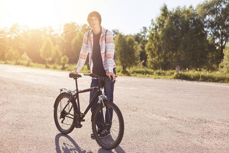 时尚潮人家伙站在他的自行车旁边同时休息后长途旅行，在照相机摆姿势，享受新鲜的空气和美丽的自然。男性青少年在周末期间在他的自行车上