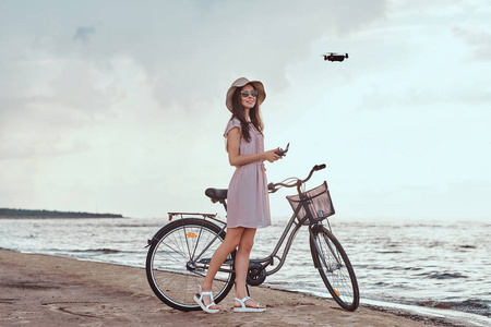 迷人的女孩在太阳镜和帽子穿着礼服享受在海滩上的假期, 玩 quadcopter