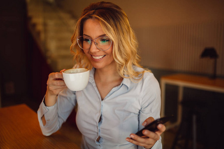 戴眼镜的年轻女子手持智能手机, 在咖啡歇的咖啡馆喝咖啡。
