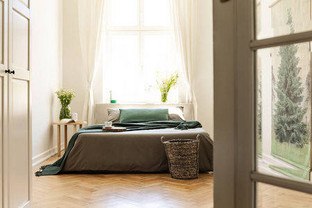 绿色毯子在灰色床在极小的卧室内部与花和窗口。真实照片