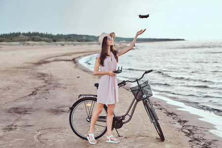 迷人的女孩在太阳镜和帽子穿着礼服享受在海滩上的假期, 玩 quadcopter