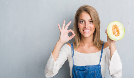美丽的年轻女子在垃圾灰墙上举行新鲜哈密瓜做 ok 标志与手指, 优秀的符号