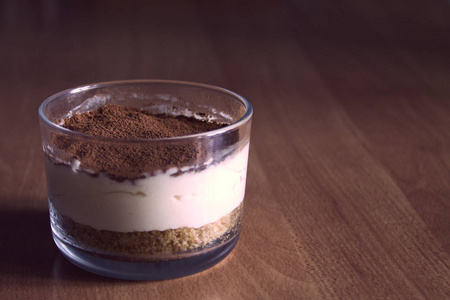在玻璃，装饰着咖啡屑，在夏天的早晨中的模糊背景上的木桌上的牛奶芝士蛋糕
