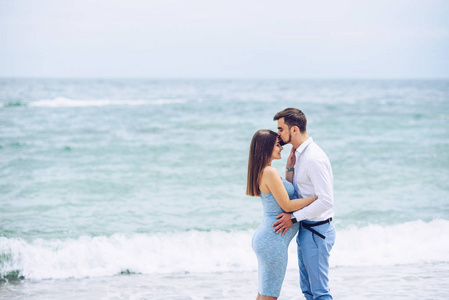 勇敢英俊的男人，在一件白衬衫和蓝色长裤的前额亲吻她美丽的怀孕的妻子在一件蓝色的连衣裙，背景下的岩石和海