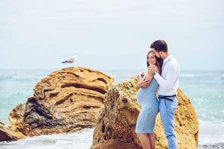 一个美丽的怀孕女人，穿一件蓝色的衣服和她勇敢和英俊的丈夫，在衬衫和蓝色的裤子靠彼此与海鸥海岩石的背景