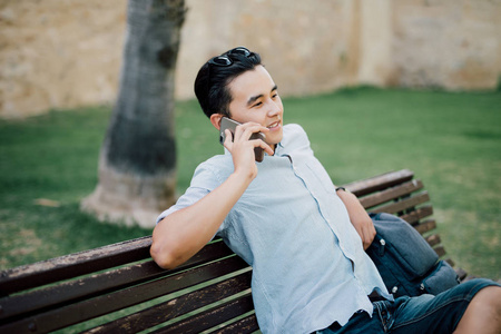 亚洲年轻英俊男子谈电话坐在公园长凳上户外的肖像