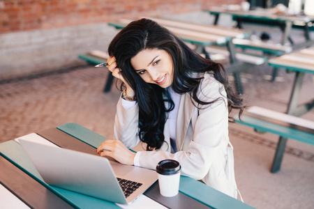 可爱的女企业家与暗长长的头发，在她的手坐在户外使用笔记本电脑她喝咖啡尝试的工作，使项目握笔。可爱的黑发女性工作