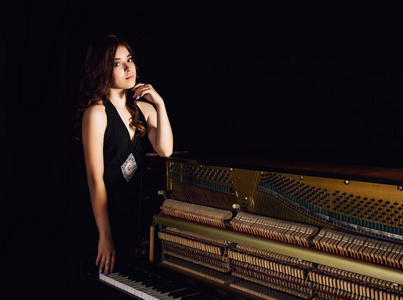 美丽的年轻女子，在开放式回站神经上深色背景的钢琴黑色连衣裙