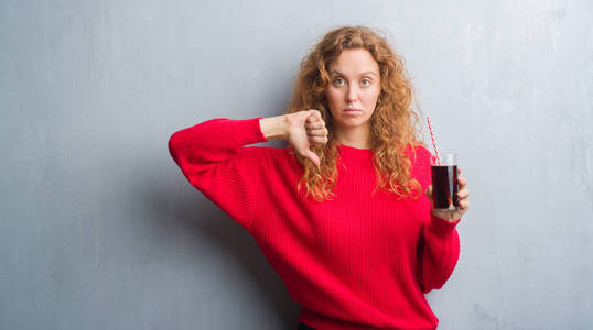 灰色垃圾墙上的年轻红头发的女人喝苏打茶点与愤怒的脸, 消极的迹象显示不喜欢拇指向下, 拒绝概念
