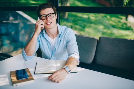 成功的白种人的肖像在光学眼镜视力矫正坐在桌子上的记事本, 并呼吁智能手机设备。积极的年轻人在电话中与接线员交谈