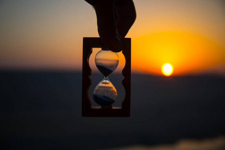 生命时间流逝的概念.手持沙漏与日落天空背景.选择性聚焦照片