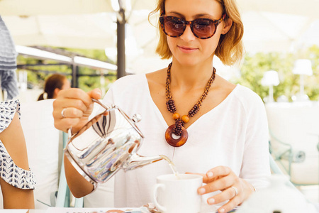 时尚的金发女郎把茶倒入杯子里, 在户外咖啡馆里坐着茶壶