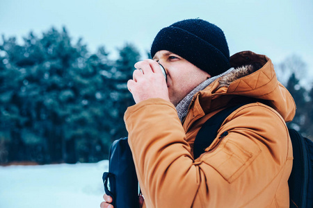 一个年轻男子在红色外套，站在雪地上，抱着一个保温瓶和一杯茶一个背包。冬季森林