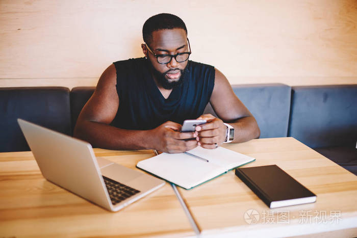 严重的非洲裔美国时髦的家伙在眼镜检查智能手机的通知, 同时学习与技术在咖啡馆内, 深色皮肤男性自由职业者使用手机检查邮件