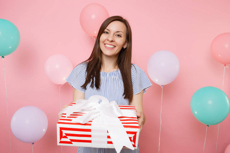 有吸引力的微笑的年轻妇女的肖像在蓝色礼服举行红色盒与礼物在柔和的粉红色背景与五颜六色的空气气球。生日聚会, 人的真诚情感理念