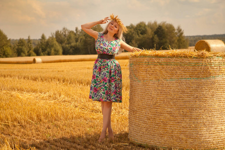 美丽的农村妇女，穿着花裙子和小麦冠站附近干圆秸秆干草堆黄色字段中