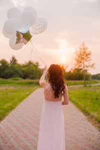 快乐美丽的女人在粉红色的长裙散步与空气气球在绿色公园在日出的夏天天