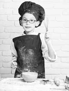 可爱的小男孩戴着眼镜，站在旁边用擀面杖表并举行厨房扫和白砖的背景墙上的绿松石碗煮红色均匀的厨师和帽子上沾满泪水的脸面粉
