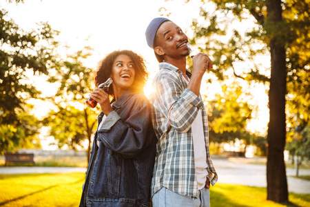 一个欢快的年轻非洲夫妇的肖像持有汽水瓶与碳酸饮料, 而站在一起在公园