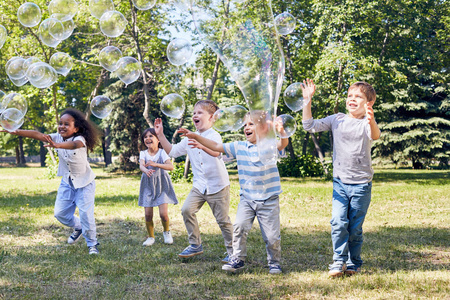 多民族的快乐的孩子们在绿色的公共公园聚会时捕捉大的肥皂泡