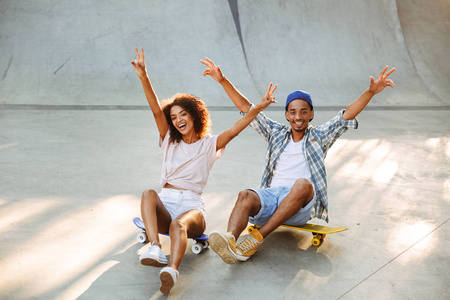 一个快乐的年轻非洲夫妇的肖像坐在一起的滑板公园与手伸出