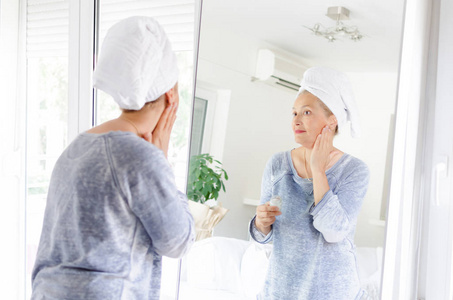 美丽的资深妇女在镜子前面使用抗衰老的奶油在家