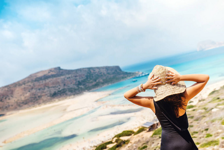 美丽的女人身穿一件泳衣和草帽, 从悬崖上看海。享受夏日和无忧无虑假期的妇女