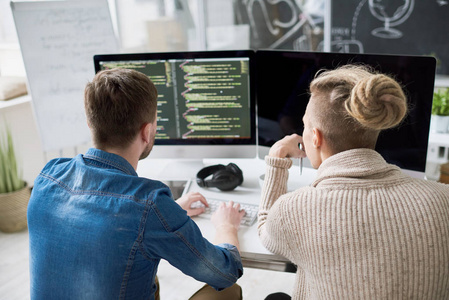 后视图的两个当代节目开发商编写代码坐在办公桌上与计算机协作, 在现代办公室的启动项目, 复制空间