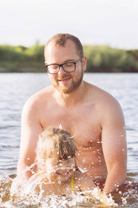 一个年轻的大胡子父亲, 戴着眼镜, 有个金发碧眼的小女儿在阳光明媚的夏日里在河里泼水。