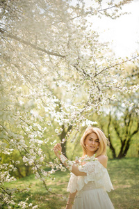 在盛开的樱桃树附近的白色礼服的幸福金发女郎的垂直浪漫肖像