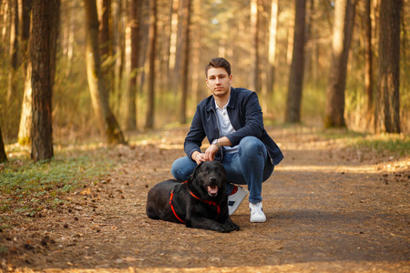 时尚的年轻人与黑狗在森林中日落照片
