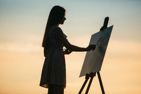 一个金发女郎的剪影。在油漆的帮助下, 女士在画布上画一幅油画。一个木画架保持图片。夏天是晴天, 日落