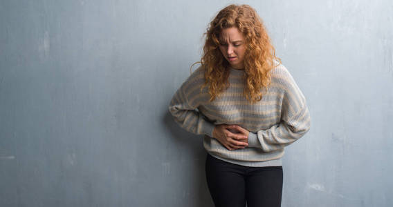 年轻的红头发女人在灰色的垃圾墙上, 手放在肚子上, 因为消化不良