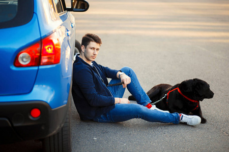 英俊的年轻人坐在一辆小汽车旁边, 靠着一只狗休息。和朋友一起旅行