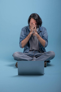 年轻的大胡子男子坐在地板上使用计算机在蓝色背景。混乱的亚洲年轻的时髦使用膝上型电脑, 全身射击。年轻一代时髦的工作理念